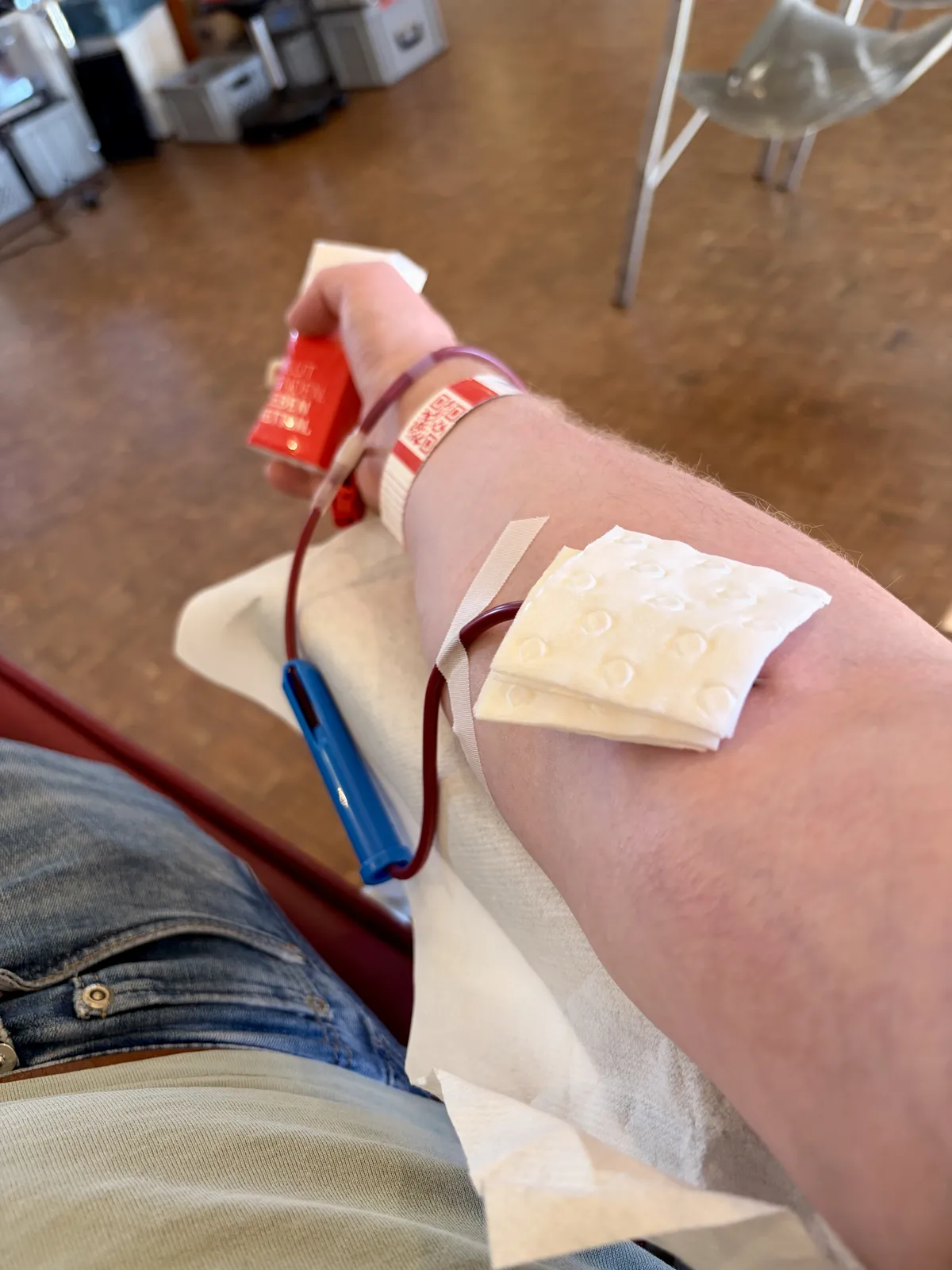 Ein Arm mit einem Verband und einer Blutabnahme. Im Hintergrund sind weitere medizinische Geräte und eine Wartebereich sichtbar.