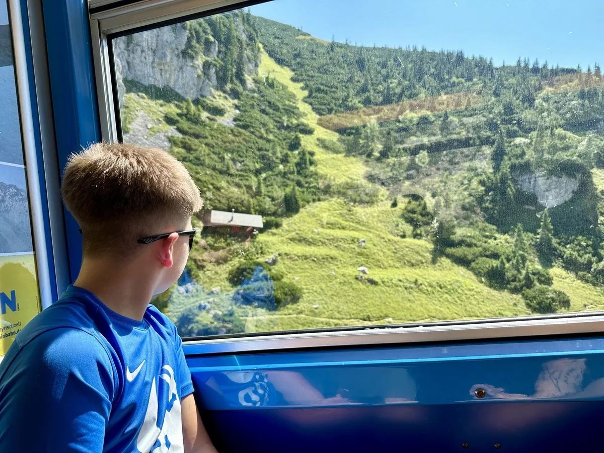 Ein Junge im blauen Trikot schaut aus dem Fenster eines blauen Zuges auf eine malerische Berglandschaft mit grünen Wiesen und Bäumen.