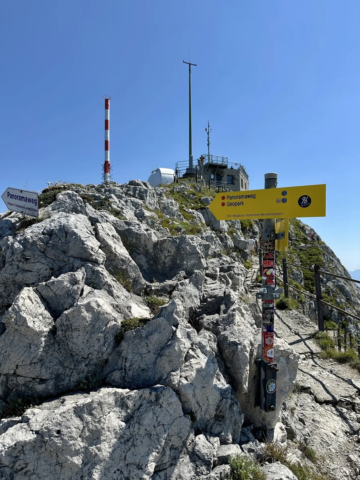Der felsige Gipfel des Wendelstein mit Wegweisern und dem Sendeturm vor einem klaren blauen Himmel.