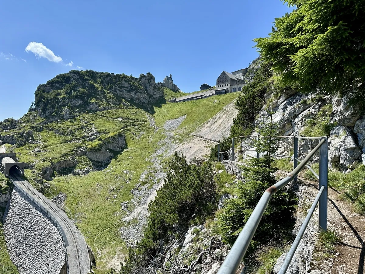 Eine malerische Berglandschaft, mit einer Eisenbahnlinie, die in einen Tunnel führt, und einem Gebäude, das auf einem Hügel thront, umgeben von grüner Vegetation.