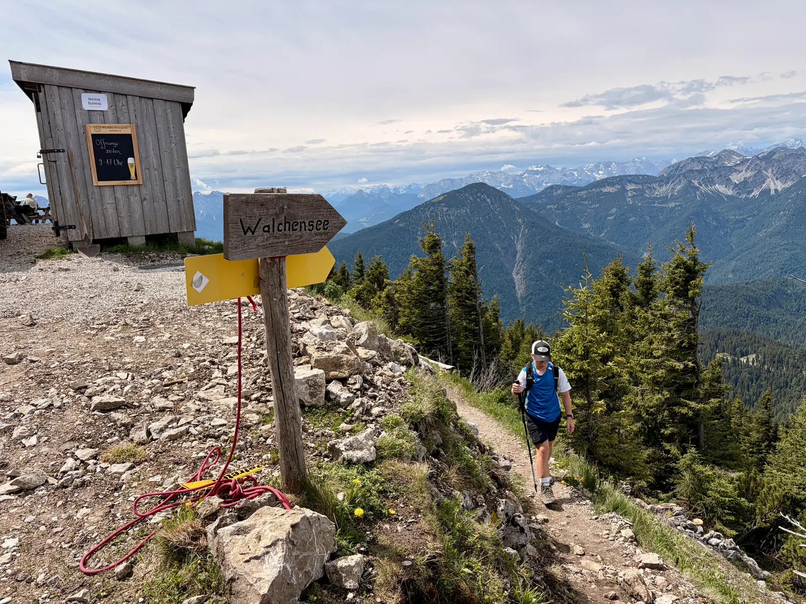 Ein Wanderer mit Wanderstöcken in der Hand kommt einen schmalen Pfad neben der Hütte auf dem Heimgarten entlang.