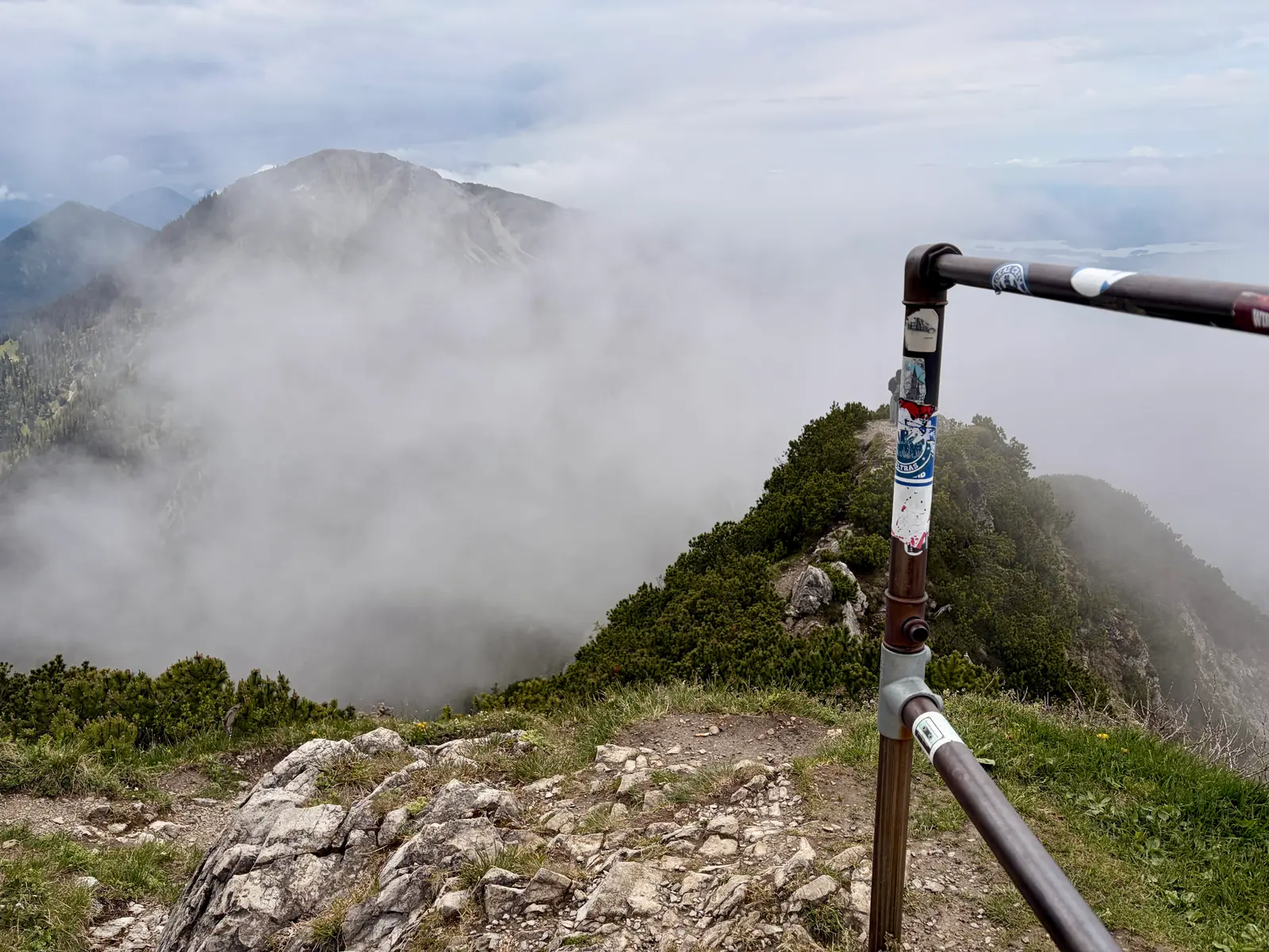 Nebel zieht über den felsigen Grat zwischen Heimgarten und Herzogstand, ein Geländer markiert den schmalen Weg. Die umliegenden Berggipfel sind nur schemenhaft durch die Wolken zu sehen.