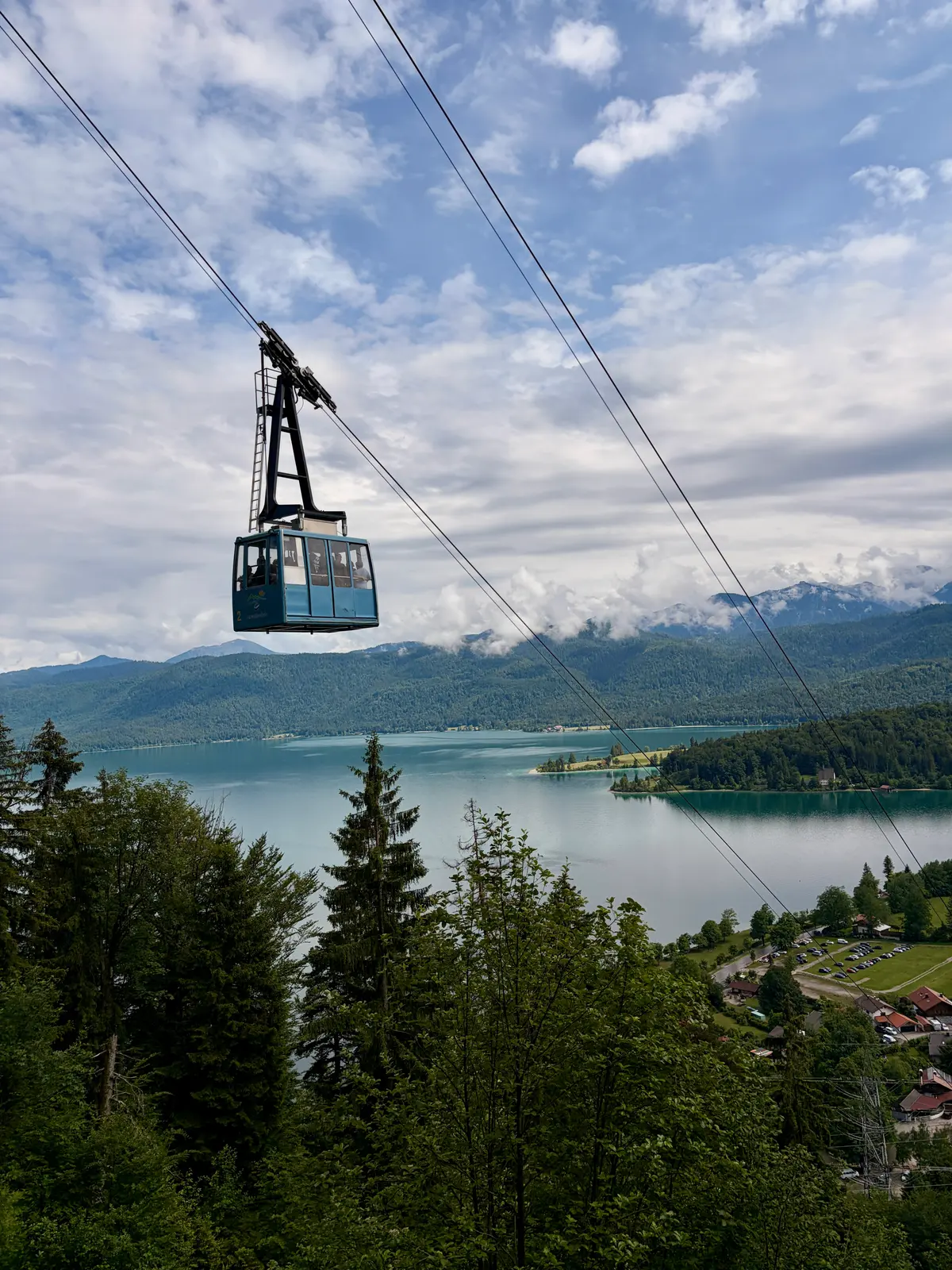 Eine Gondel der Herzogstandbahn schwebt über dem Walchensee, dahinter bewaldete Hänge und wolkenverhangene Berge. Im Vordergrund ragen Tannen in den Blick auf den türkisfarbenen See.