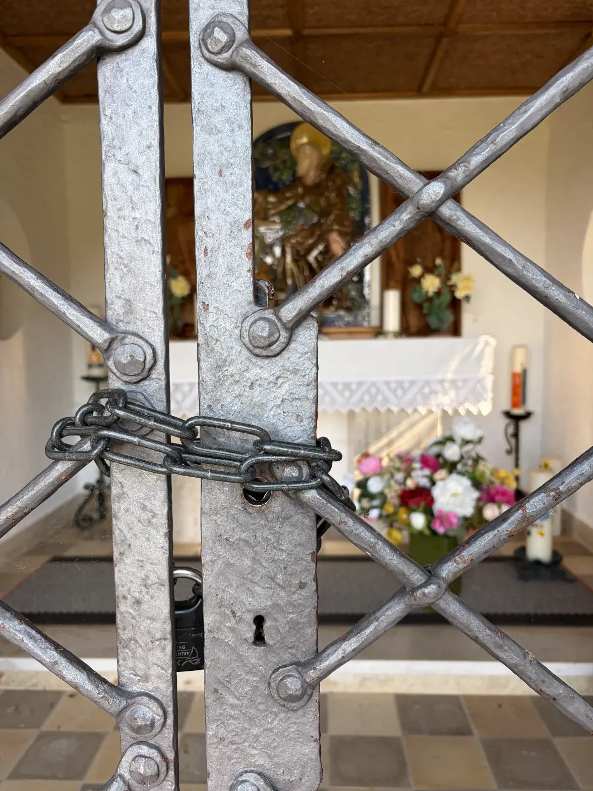 Metallgitter mit Kette und Schloss vor dem Eingang der St.-Michaels-Kapelle an der Schwarzenkopfhütte; im Hintergrund sind Altar, Blumen und Kerzen zu sehen.