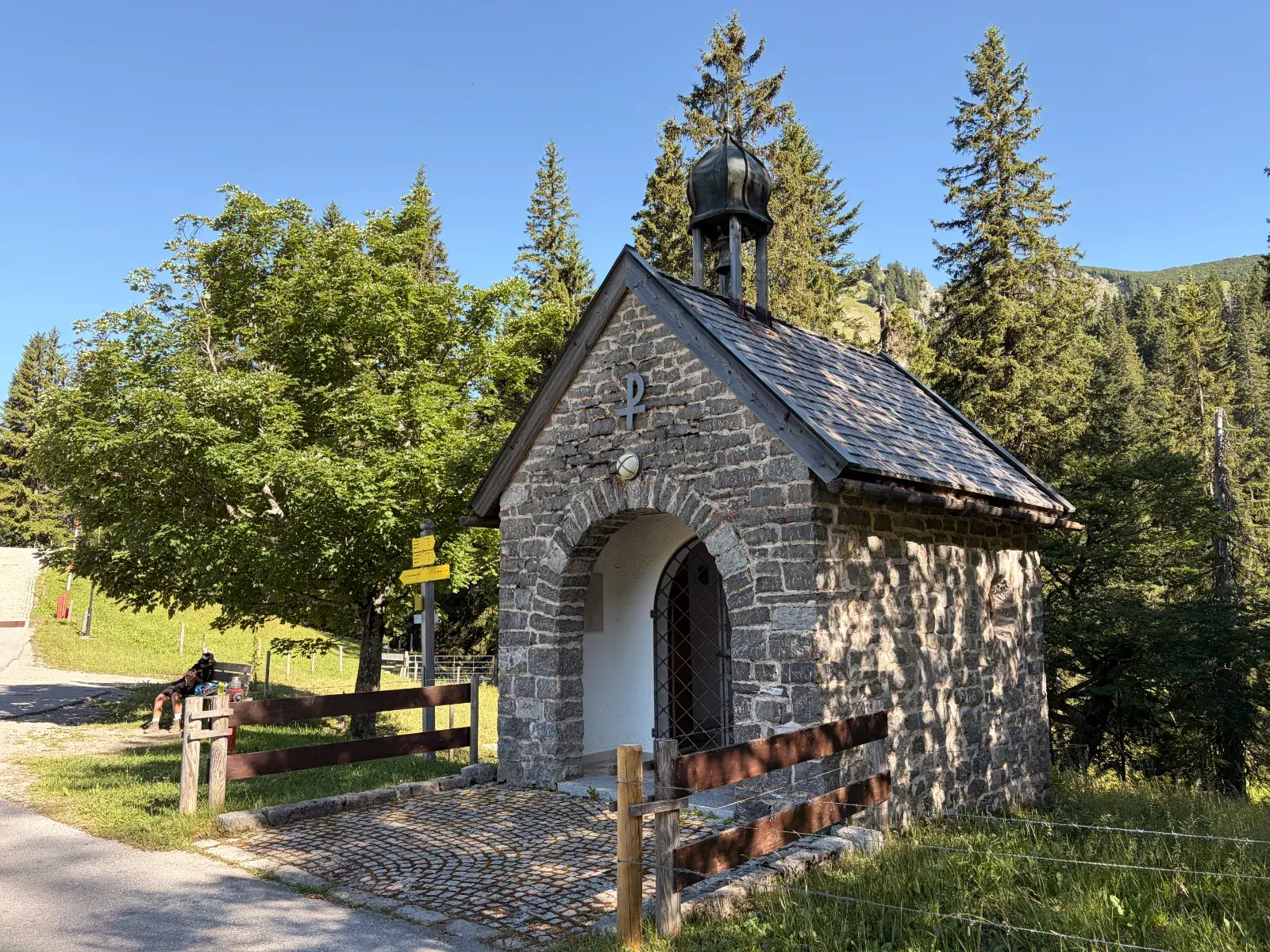 Kleine steinerne St.-Michaels-Kapelle an der Schwarzenkopfhütte bei Spitzingsee, umgeben von Bäumen und Holzzaun. Im Hintergrund steigen bewaldete Hänge unter klarem Himmel an.
