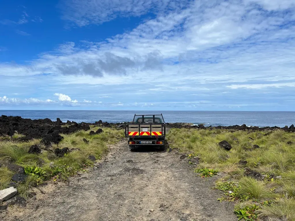 Ein Lieferwagen steht auf einem Schotterweg zwischen schwarzem Vulkangestein und Gras, dahinter der einsame Strand und das Meer der Azoren.