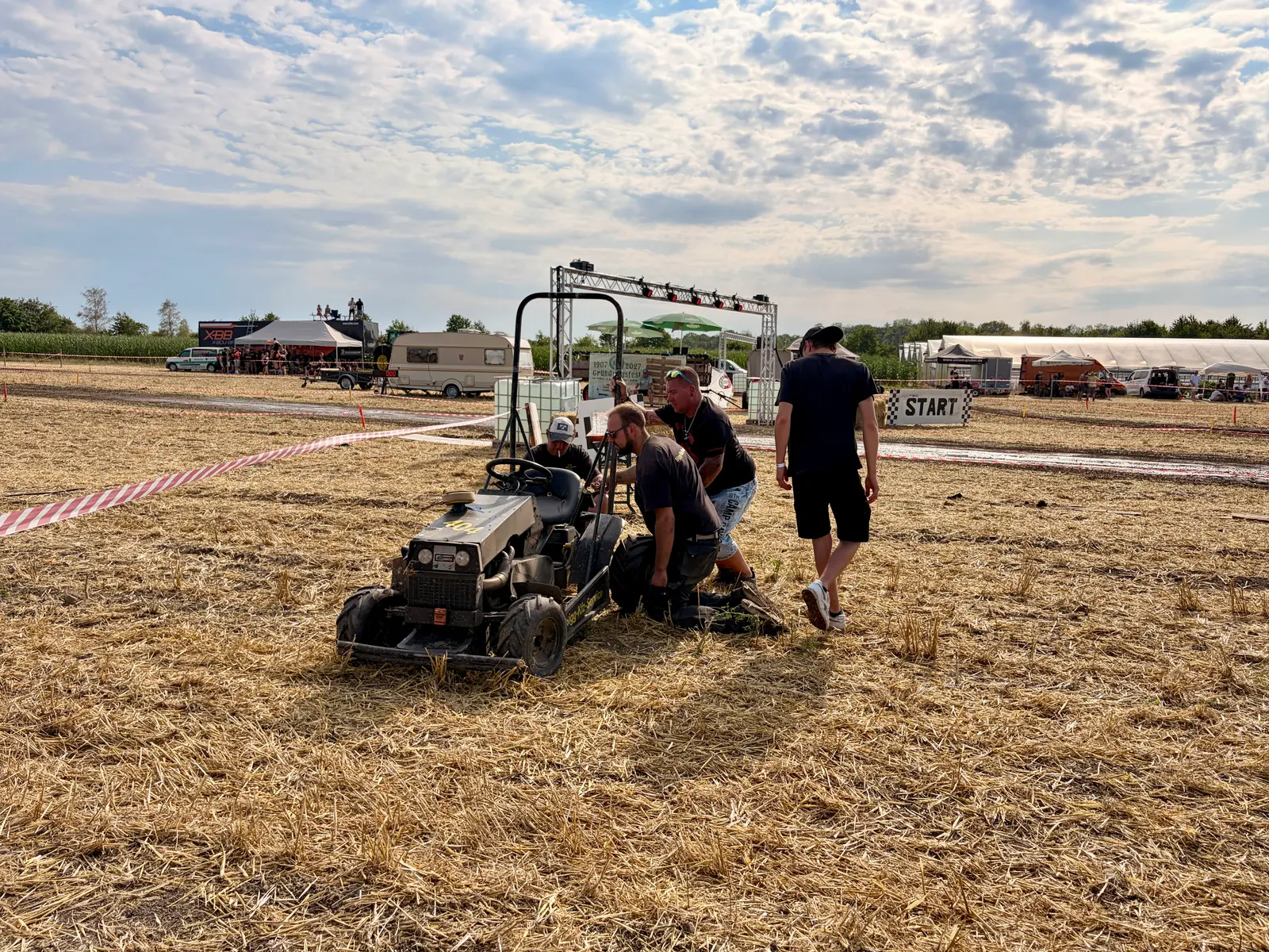 Mehrere Personen reparieren einen umgebauten Rasenmäher auf einem staubigen Stoppelfeld nahe der Startlinie. Im Hintergrund stehen Zelte, Fahrzeuge und Absperrband unter einem wolkigen Himmel.
