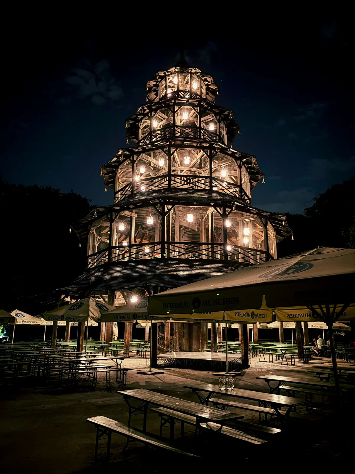 Beleuchteter chinesischer Turm im Englischen Garten bei Nacht, umgeben von dunklen Bäumen und Biergarten-Schirmen. Im Vordergrund stehen leere Bierbänke und Biertische.