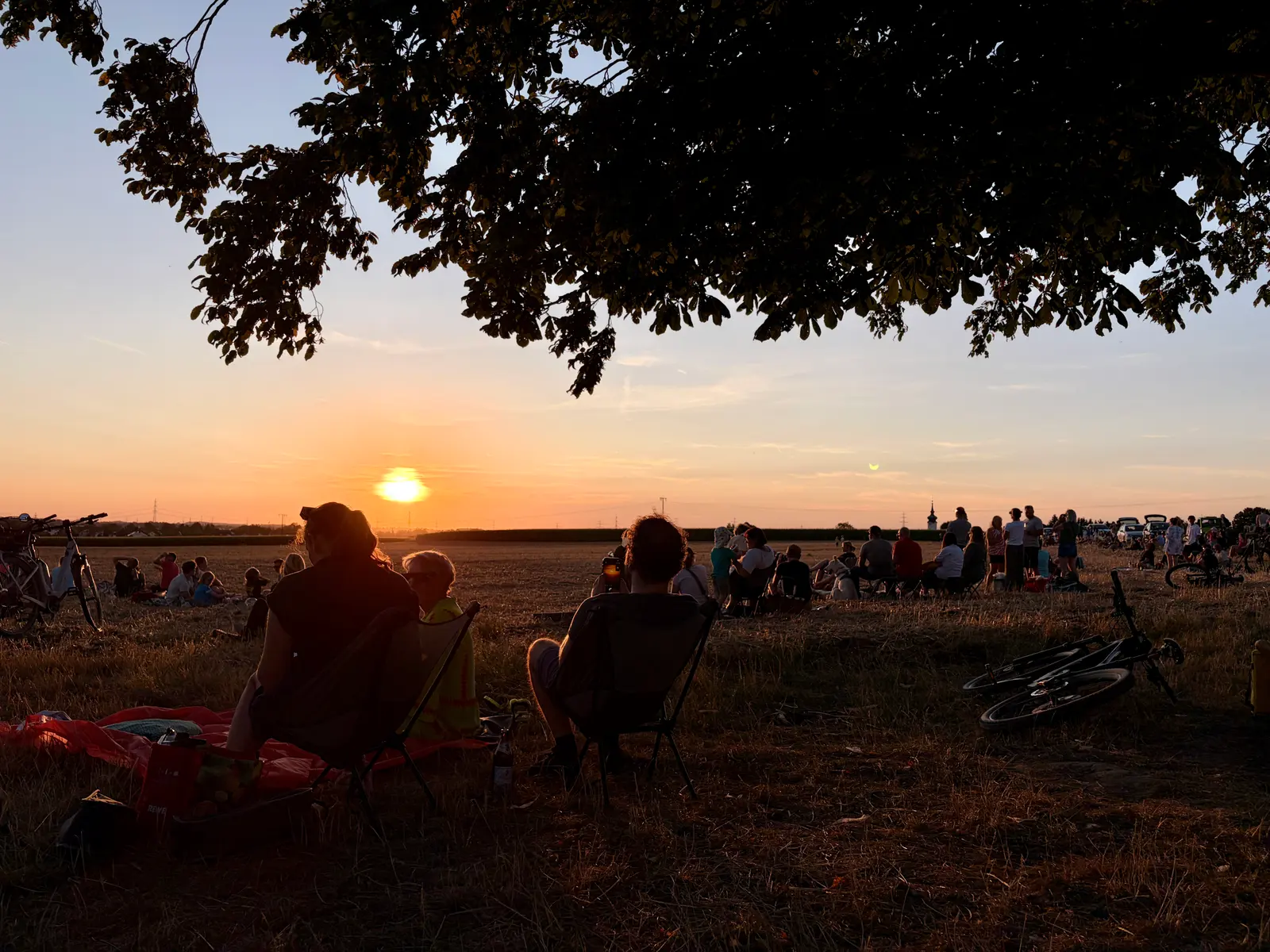 Menschen sitzen bei der Sonnenfinsternis auf einer Wiese unter einem Baum, einige schauen Richtung Horizont. Fahrräder liegen und stehen am Rand, im Hintergrund sind weitere Gruppen zu sehen.