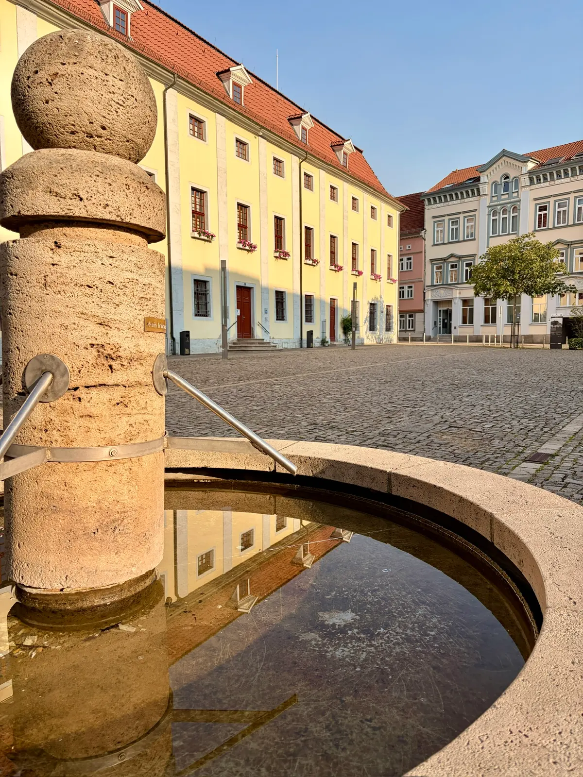 Historischer Platz in Mühlhausen mit Kopfsteinpflaster, gelbem Gebäude und roten Dächern im warmen Morgenlicht. Im Vordergrund spiegelt sich die Häuserzeile in einem kleinen runden Wasserbecken neben einer steinernen Säule.