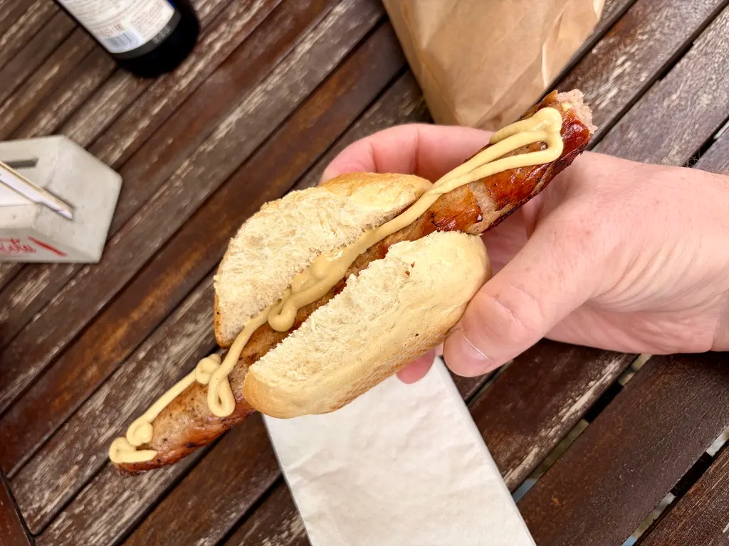 Eine Hand hält eine Bratwurst im Brötchen mit Senf über einem Holztisch. Im Hintergrund liegen eine Serviette, eine Papiertüte und eine Flasche.