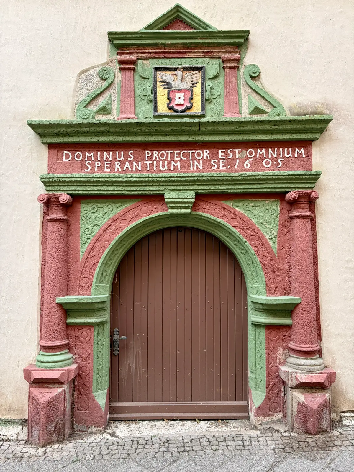 Historisches Portal mit brauner Holztür, rot-grüner Steinverzierung und lateinischer Inschrift an einer hellen Fassade in Mühlhausen.